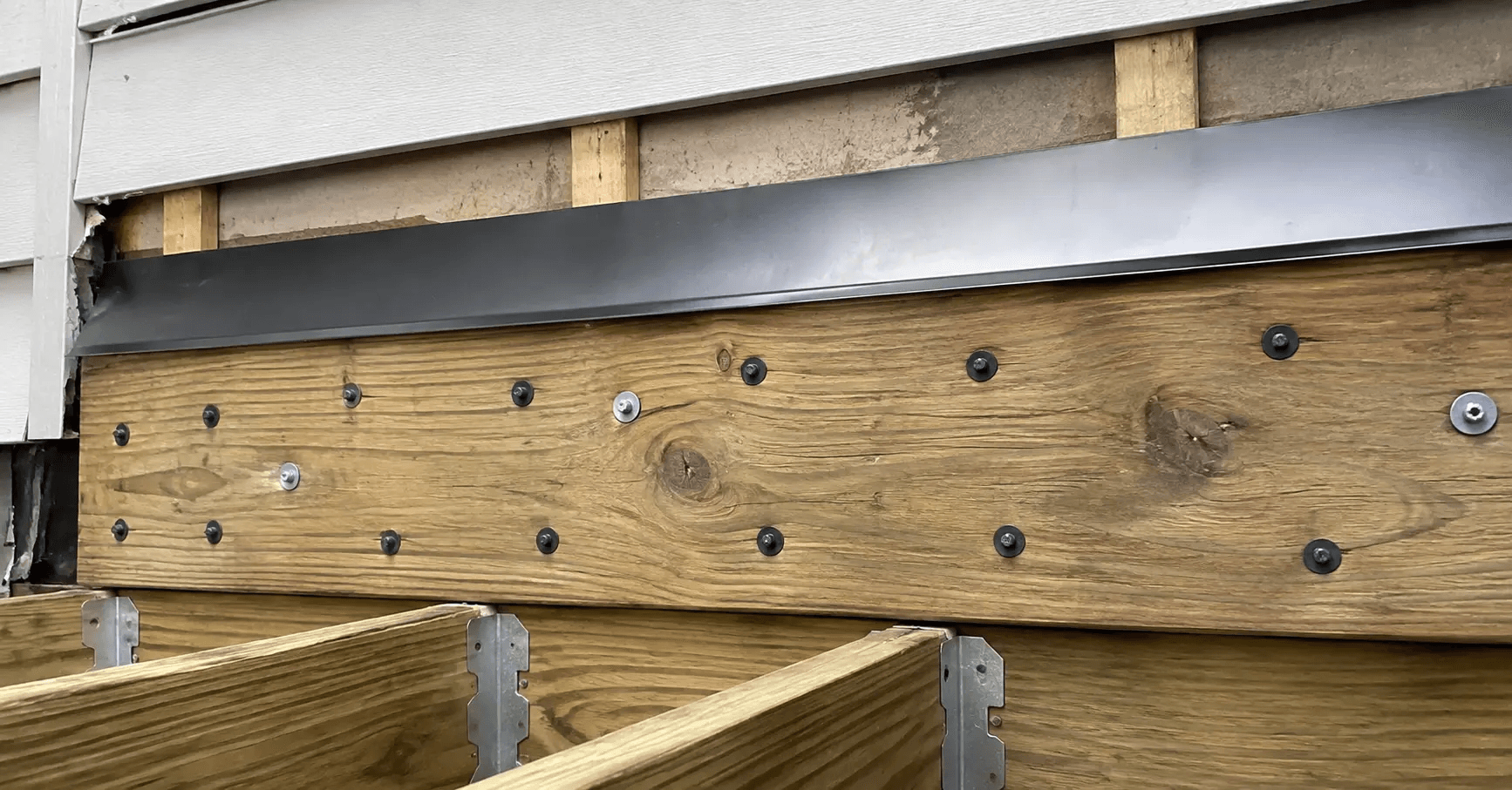 Deck ledger board fastened to an exposed rim joist with cap flashing installed above it, siding cut back